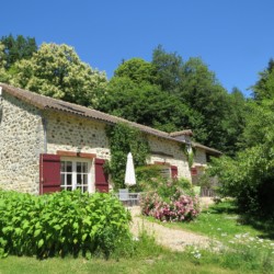 Rustic stone cottage surrounded by lush greenery, with blooming flowers and a birch tree (bouleau) in the foreground, under a clear blue sky.