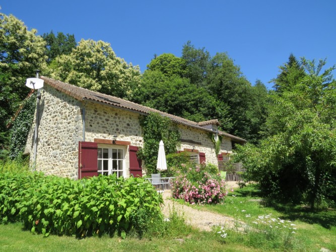 Rustic stone cottage surrounded by lush greenery, with blooming flowers and a birch tree (bouleau) in the foreground, under a clear blue sky.
