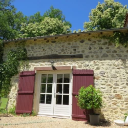Quaint stone cottage with wooden shutters and a small potted tree in front, named Châtaignier, a gite surrounded by greenery under a clear blue sky.