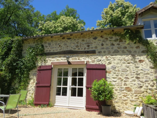 Quaint stone cottage with wooden shutters and a small potted tree in front, named Châtaignier, a gite surrounded by greenery under a clear blue sky.